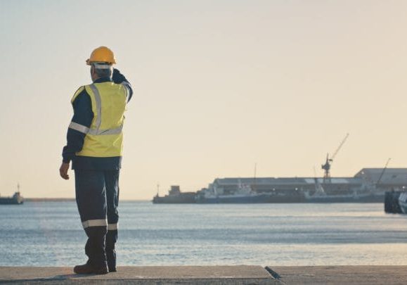 Shipping, freight and logistics with a supervisor standing on the dock in a harbor, looking at the view and waiting for a delivery or shipment. Safety and control in the import and export industry