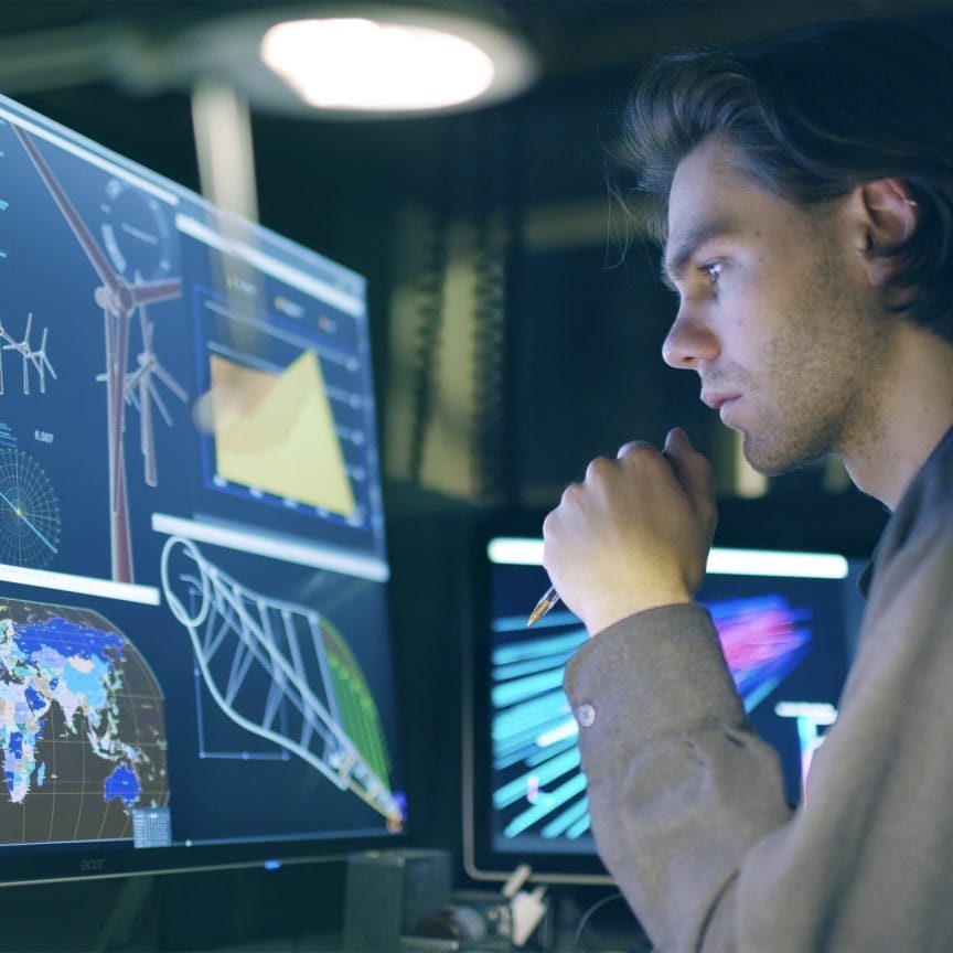 Stock image of a young, long haired man,  working at night in front of a large computer screen showing designs for a wind turbine (wind farm)
