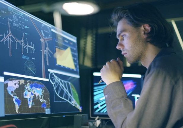 Stock image of a young, long haired man,  working at night in front of a large computer screen showing designs for a wind turbine (wind farm)